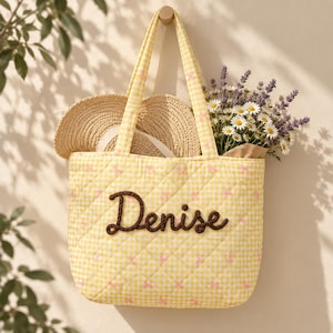 May include: A quilted yellow and white gingham tote bag featuring the name Denise embroidered in dark brown yarn on the front. The bag contains a straw hat and a bouquet of white daisies and purple lavender flowers, hanging from a wooden hook.