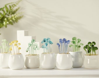 May include: Eight white ceramic pots with different colored glass plant sculptures. The plants are in shades of green, blue, and yellow.