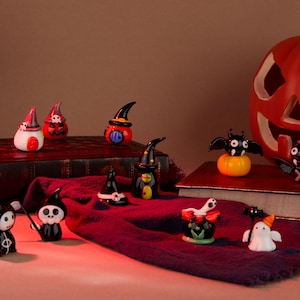 以下が含まれることがあります： 幽霊、コウモリ、魔女、カボチャなど、ハロウィーンをテーマにしたガラス製の置物コレクション。置物は赤い布と本の上に置かれています。背景には、顔の彫刻が施された大きなオレンジ色のカボチャがあります。