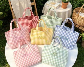 May include: A collection of six small tote bags in various gingham patterns including red, light pink, yellow, light green, and light blue. Each bag features two handles and a name embroidered in cursive script on the front center of the fabric.