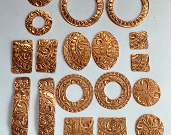 May include: A collection of 16 copper-colored metal charms with various designs, including circles, squares, and floral patterns. Some charms have a raised edge, while others are flat. The charms are arranged on a white surface.
