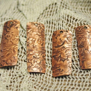 May include: Four copper-colored metal tags with an ornate design. The tags are rectangular and have a slightly curved shape. They are arranged in a row on a white crocheted surface.