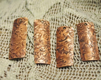 May include: Four copper-colored metal tags with an ornate design. The tags are rectangular and have a slightly curved shape. They are arranged in a row on a white crocheted surface.