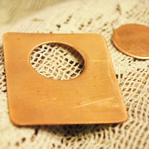 May include: A copper square with a round hole cut out of the center. The copper square is sitting on a white crocheted surface.
