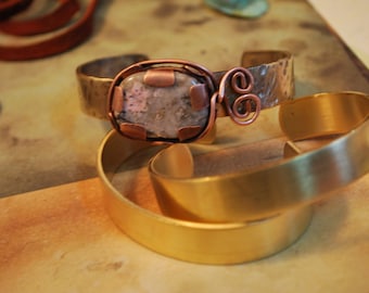 May include: A copper bracelet with a hammered finish and a gray stone set in a copper bezel. The bracelet has a copper wire accent in the shape of the letter "S".