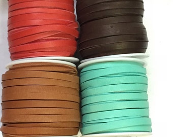 May include: Four spools of flat leather cord in various colors. The colors include red, dark brown, tan, and teal. Each spool is neatly wound on a white plastic bobbin, ready for crafting or jewelry making.