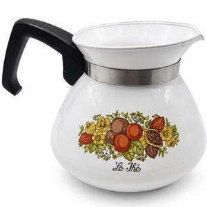 May include: A white enamel teapot with a black handle and a silver-colored metal band around the neck. The body features a colorful botanical illustration of flowers, leaves, and fruit, with the black cursive text Le Thé printed below the design.