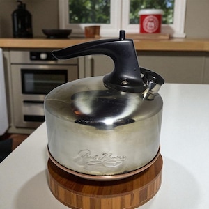 May include: A stainless steel kettle with a black handle and spout sits on a wooden trivet. The kettle has a shiny, reflective surface and a copper-colored ring at the base. The trivet is made of wood and has a circular design.