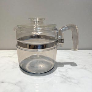 May include: A clear glass coffee pot with a silver metal band around the middle. The pot has a clear handle and a glass lid with a knob. The pot is sitting on a white marble surface.