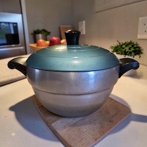 May include: Vintage aluminum cooking pot with a teal lid and black handle. The pot sits on a wooden cutting board. The lid has a black knob. The pot is a kitchenware item.