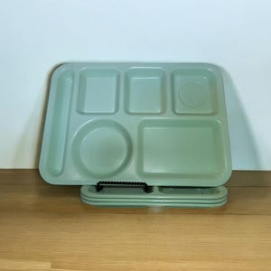 May include: A stack of light sage green rectangular cafeteria trays, each featuring one long narrow compartment, two smaller rectangular sections, one circular indentation, and one large rectangular area for food service.