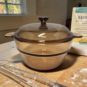 May include: A round, amber-colored glass casserole dish with a matching lid. The pot features integrated side handles and a central knob on the lid. The glass is translucent, showing a smooth, curved design with a dark horizontal band around the center.