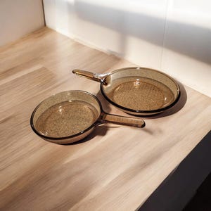May include: Two round, transparent amber-colored glass skillets with integrated handles. The pans feature a smooth, polished finish and a shallow depth, showcasing a consistent brownish tint throughout the glass material of both cooking vessels.