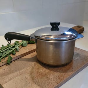 May include: A stainless steel cooking pot with a lid and a black handle. The pot sits on a wooden cutting board with fresh green herbs. The lid has a black knob.