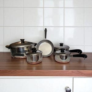 May include: A set of stainless steel cookware with copper-colored bottoms and black handles. The set includes pots, pans, and a frying pan, all arranged on a wooden surface against a white tiled background.