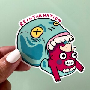 Könnte beinhalten: Ein Aufkleber mit einem blauen Monster mit einem großen Maul und einem roten Monster mit einer Krone im Maul. Der Aufkleber trägt oben die Aufschrift "REINTARNATION".