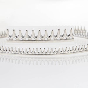 Puede incluir: Un accesorio para el cabello de metal plateado en forma de peine. El accesorio tiene forma circular con una pieza rectangular más pequeña del mismo diseño. Los dientes del peine están espaciados uniformemente y curvados hacia arriba.