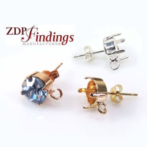 May include: Two pairs of earring settings, one gold and one silver. Each setting has a round bezel for a stone and a post for an earring. The gold setting has a blue stone in the bezel.