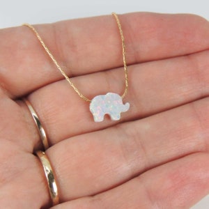 May include: A gold-colored necklace featuring a small, white opal elephant pendant. The elephant has a textured surface and is suspended from a delicate chain. The pendant is a light, iridescent color.