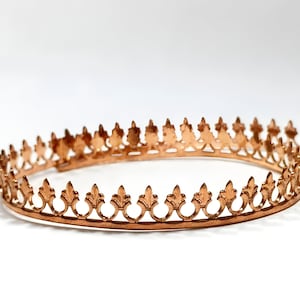 May include: A copper-colored crown with a repeating fleur-de-lis design. The crown is circular and has a series of pointed elements along the top edge. The metal has a warm, reflective sheen, suggesting it is made of copper or a copper alloy.