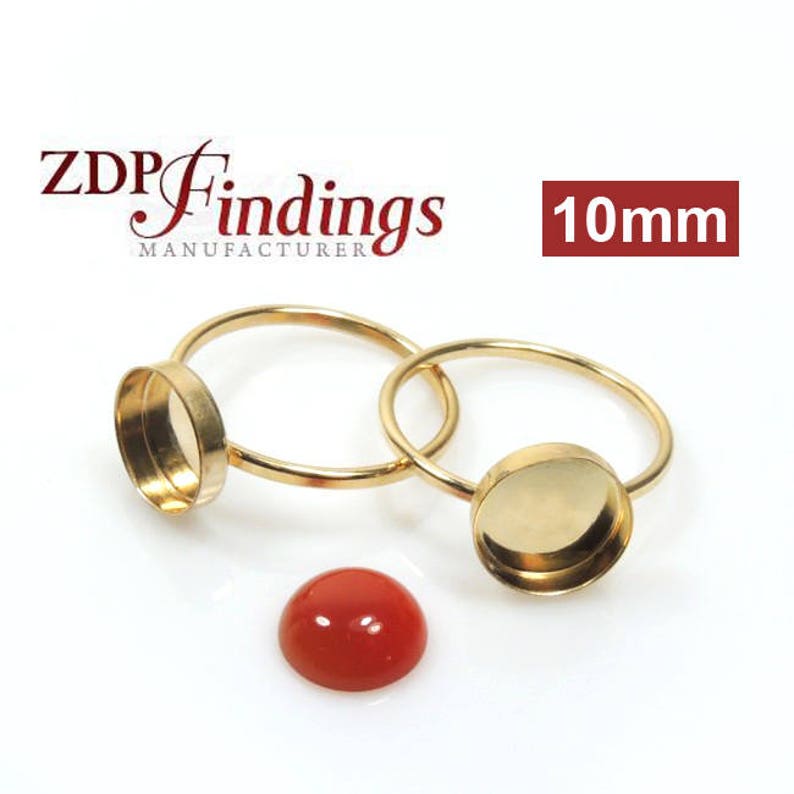 May include: Two gold-toned metal ring settings with 10mm round bezels. The bezels are empty and ready to be filled with a cabochon stone. The text "ZDP Findings Manufacturer" and "10mm" are visible in the image. A red cabochon stone is also shown.