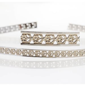 May include: A silver headband with a delicate floral design. The headband is made of metal and has a gold-toned floral pattern.