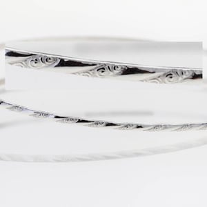 May include: A silver bangle bracelet with a delicate, etched design. The bracelet is thin and has a simple, elegant style.