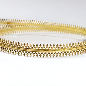 Op de afbeelding: Een gouden metalen kroon met een delicaat, ingewikkeld ontwerp. De kroon is gemaakt van een dunne, opengewerkte band met een herhalend patroon van puntige bogen.