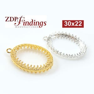 Könnte beinhalten: Zwei ovale Metall-Anhänger-Einstellungen, eine goldene und eine silberne, mit einem dekorativen kronenartigen Rand. Die Einstellungen sind 30x22 mm groß.