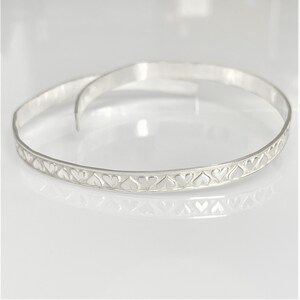 May include: Two silver bangles. One bangle is open, and the other is closed. Both have a row of heart-shaped cutouts along the outer edge. The bangles are displayed on a white reflective surface.