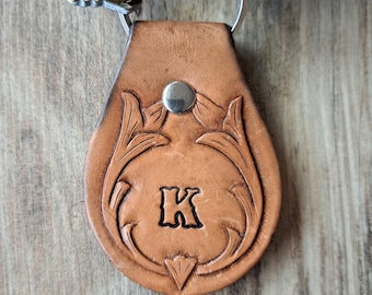 May include: A brown leather keychain with a silver metal snap and a metal keyring. The keychain features a carved floral design and the letter "K". The keychain is attached to a set of keys.