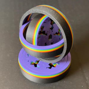 May include: A 3D-printed sculpture featuring a black ring with a rainbow stripe, intersecting a purple base with flower cutouts. The sculpture has a rainbow-colored ring inside the black ring. The base is also purple with a rainbow stripe.