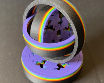 Puede incluir: Una escultura impresa en 3D con un anillo negro y una franja arcoíris, que cruza una base púrpura con recortes de flores. La escultura tiene un anillo de color arcoíris dentro del anillo negro. La base también es púrpura con una franja arcoíris.