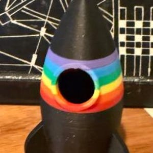 May include: A black rocket-shaped decorative item with a rainbow-colored band around the middle. The rocket has a cone-shaped top and two black fins. The item has a circular opening in the middle.