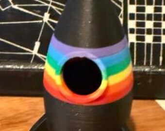 Puede incluir: Un objeto decorativo negro con forma de cohete con una banda de arcoíris en el medio. El cohete tiene una parte superior en forma de cono y dos aletas negras. Tiene una abertura circular en el medio.