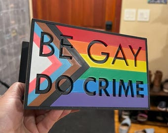 Puede incluir: Un letrero rectangular con el texto "BE GAY DO CRIME" en letras negras recortadas. El letrero presenta el diseño de la bandera del Orgullo Progresista, con un arcoíris y colores adicionales. El letrero está sostenido en una mano.