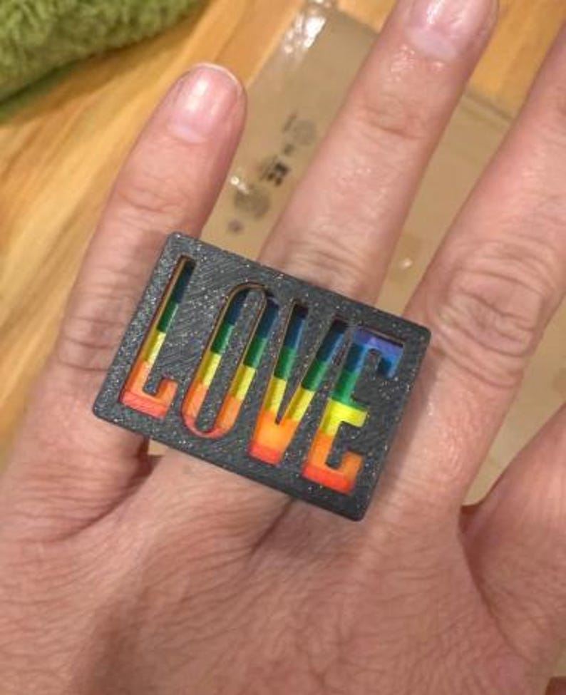 Puede incluir: Un anillo rectangular gris oscuro con la palabra "LOVE" recortada, que revela un degradado arco&iacute;ris. Las letras del anillo est&aacute;n rellenas de rojo, naranja, amarillo, verde y azul. El anillo tiene una textura brillante.