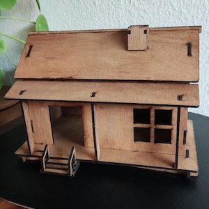 Puede incluir: Un modelo de madera de una casa pequeña con un techo marrón, una chimenea y un porche con escalones. La casa está sentada sobre una base ovalada negra.