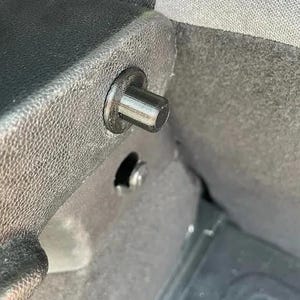 May include: Close-up of a black plastic cylindrical fastener with a wide, flat head. The fastener is inserted into a textured, dark gray surface, likely part of a vehicle's interior. A small, round hole and a metal screw are visible nearby.