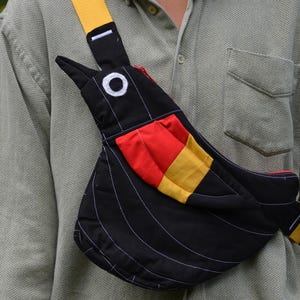 Puede incluir: Bolso bandolera negro con forma de p&aacute;jaro y correa amarilla. El bolso presenta un parche rojo y amarillo y detalles de costura blancos. El bolso se lleva sobre una camisa verde claro.