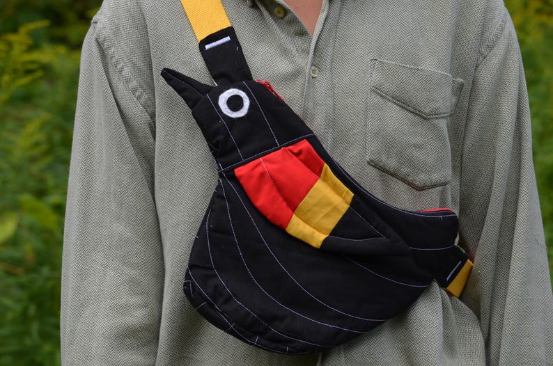 Puede incluir: Bolso bandolera negro con forma de p&aacute;jaro y correa amarilla. El bolso presenta un parche rojo y amarillo y detalles de costura blancos. El bolso se lleva sobre una camisa verde claro.