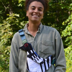 Puede incluir: Un bolso bandolera &uacute;nico con un dise&ntilde;o de p&aacute;jaro con patrones geom&eacute;tricos en negro, blanco y azul. El bolso tiene una correa azul y un detalle de ojo rojo. El fondo es follaje verde.