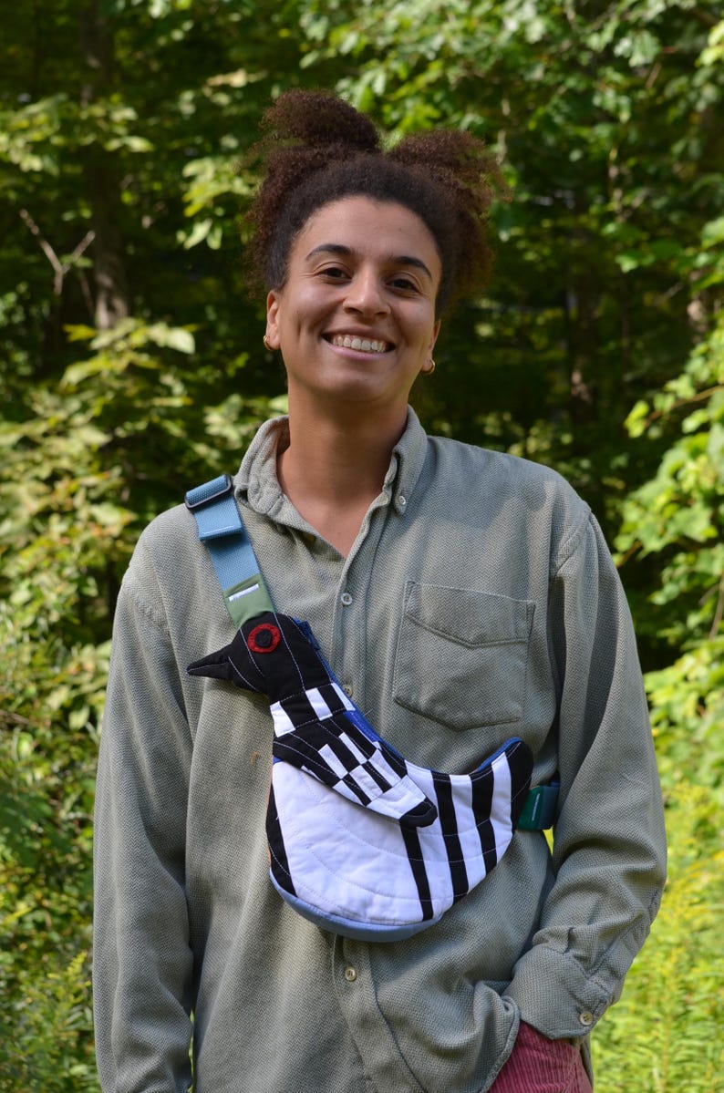 Puede incluir: Un bolso bandolera &uacute;nico con un dise&ntilde;o de p&aacute;jaro con patrones geom&eacute;tricos en negro, blanco y azul. El bolso tiene una correa azul y un detalle de ojo rojo. El fondo es follaje verde.