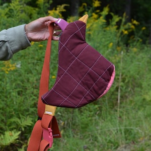 Puede incluir: Un bolso bandolera acolchado burdeos con interior rosa y un acento amarillo mostaza. El bolso tiene una correa de color &oacute;xido con una hebilla rosa. El bolso se sostiene sobre un fondo de follaje verde.