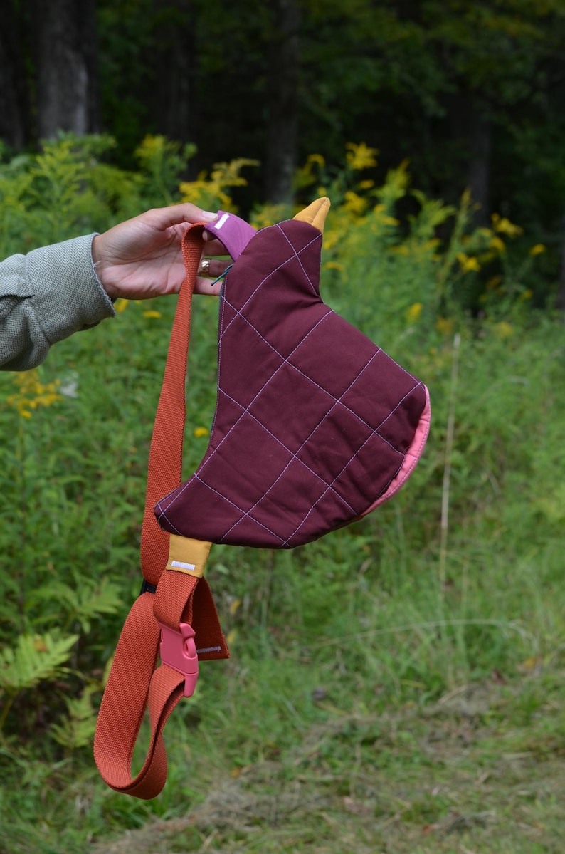 Puede incluir: Un bolso bandolera acolchado burdeos con interior rosa y un acento amarillo mostaza. El bolso tiene una correa de color &oacute;xido con una hebilla rosa. El bolso se sostiene sobre un fondo de follaje verde.