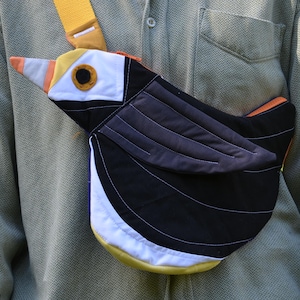 Puede incluir: Bolso bandolera con forma de p&aacute;jaro en negro, blanco, amarillo, naranja y gris. El bolso presenta una correa amarilla y un dise&ntilde;o detallado que se asemeja a un p&aacute;jaro. El bolso se lleva sobre una camisa verde.