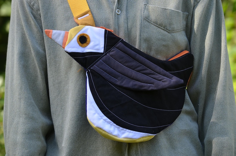 Puede incluir: Bolso bandolera con forma de p&aacute;jaro en negro, blanco, amarillo, naranja y gris. El bolso presenta una correa amarilla y un dise&ntilde;o detallado que se asemeja a un p&aacute;jaro. El bolso se lleva sobre una camisa verde.