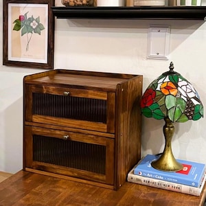 May include: A two-drawer wooden cabinet with a warm brown finish. The cabinet has a rectangular shape with two drawers featuring vertical ribbed detailing and small metal handles. A stained glass lamp with floral designs sits on top of books next to the cabinet.