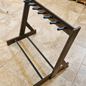 May include: A wooden wine rack with a dark brown finish. The rack has a slanted design with multiple black pegs for holding wine bottles. Two black metal bars run along the bottom for stability. The rack is designed to hold multiple bottles.