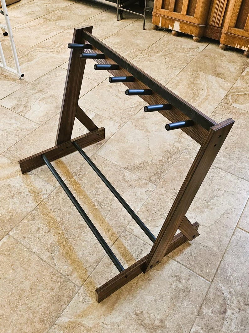 May include: A wooden wine rack with a dark brown finish. The rack has a slanted design with multiple black pegs for holding wine bottles. Two black metal bars run along the bottom for stability. The rack is designed to hold multiple bottles.